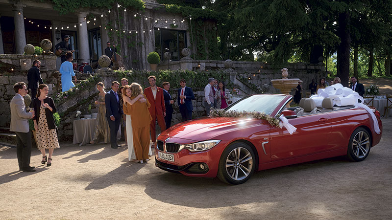 Vediamo un gruppo di invitati e una BMW rossa addobbata per gli sposi all'esterno di una villa